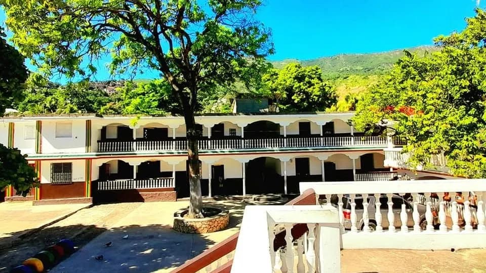 La cour de l'Institution Shalom du Cap-Haïtien, un grand arbre devant le bâtiment et les mornes au loin.